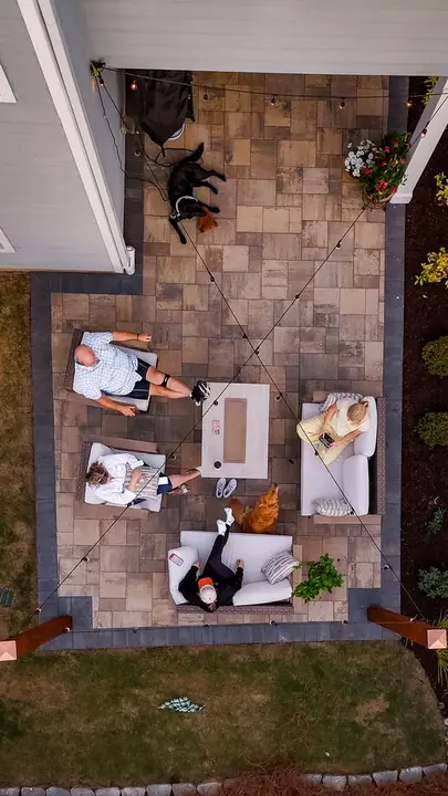 Aerial view of a paver patio with lounge seating and string lights, bordered by planting beds