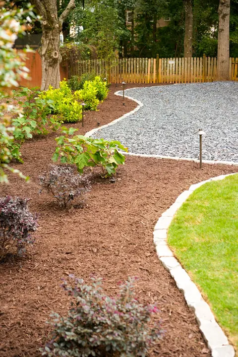 Curved mulch bed with young shrubs and stone edging beside a gravel area and wood fence
