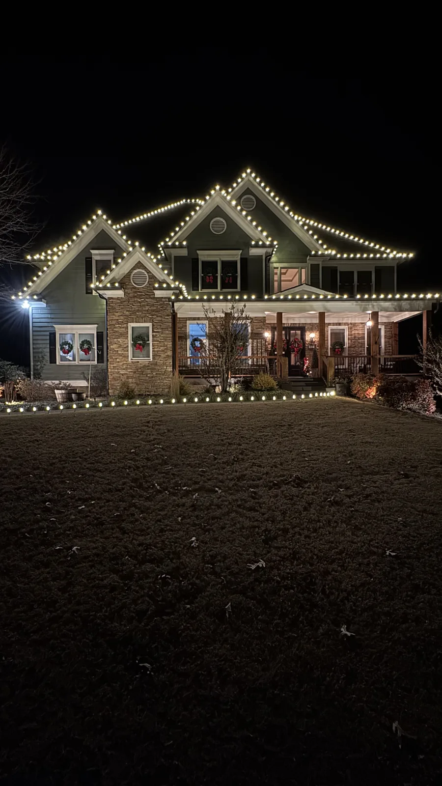 A craftsman-style home outlined in warm white roofline lights with lit wreaths, a KalKare holiday light installation at night