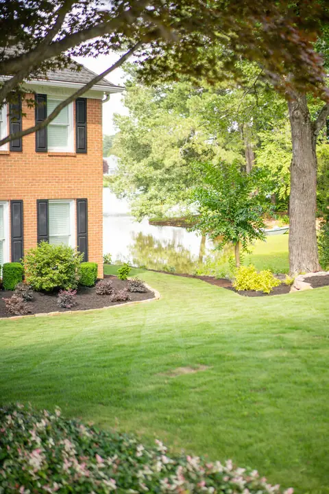 Brick lakeside home with a manicured lawn, mature shade tree, and layered planting beds
