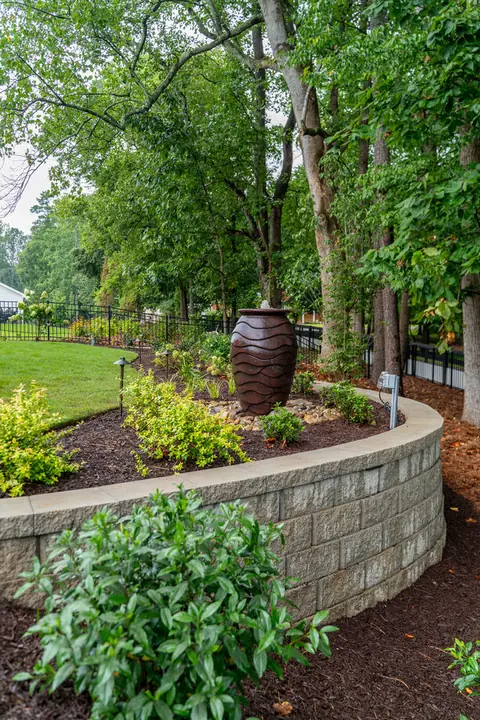 Curved stacked-stone retaining wall with a decorative urn and planting bed beneath tall shade trees