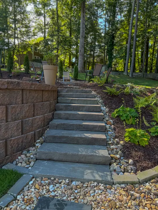 Natural stone slab steps between a retaining wall and river-rock beds leading up a wooded slope